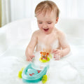 A happy baby with wet hair is sitting in a bubble bath, playing with a HAPE Tubing Pull-back boat and a tubing bear in a green float ring. The baby is surrounded by white foamy bubbles and is smiling joyfully. The background shows a white bathtub with sheer curtains.