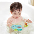 A young child with wet hair is playing in a bubble-filled bathtub. The child is holding a HAPE Tubing Pull-back boat and looking intently at a floating toy bear nearby, both toys adding to the playful atmosphere in the tub.