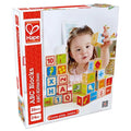 The image displays the packaging of Hape ABC Blocks by Hape. It features a young child engaging with vibrant wooden alphabet blocks that showcase letters, numbers, and pictures. The box reads "ABC Blocks," "26 pcs," and "24M+." The slogan "Love play. Learn." is also prominently shown.