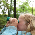 A young child with blonde hair and a blue jacket peers through the Hape Adjustable Telescope, an essential adventure accessory from the brand Hape, while standing in a lush, green forest. The background is slightly blurred, emphasizing the child's focus on bird watching and observing something in the distance.