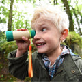 A young boy with blond hair, wearing a green jacket and plaid shirt, looks through the Hape Adjustable Telescope with excitement. He is outside in a lush, green forest, appearing thrilled and curious about his surroundings as he indulges in bird watching using this high-quality instrument from Hape.