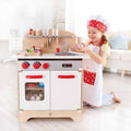 A young girl, wearing a chef's hat and apron, eagerly plays with the delightful Hape Chef Pack. She appears to be preparing toy food with a cheerful expression. The Hape kitchen set features a stovetop, oven, and sink area with children's cooking accessories hanging above.