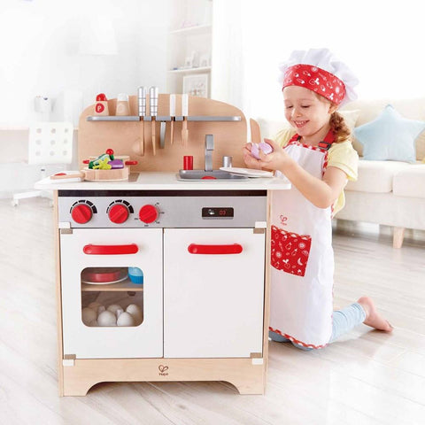 A young girl, wearing a chef's hat and apron, eagerly plays with the delightful Hape Chef Pack. She appears to be preparing toy food with a cheerful expression. The Hape kitchen set features a stovetop, oven, and sink area with children's cooking accessories hanging above.