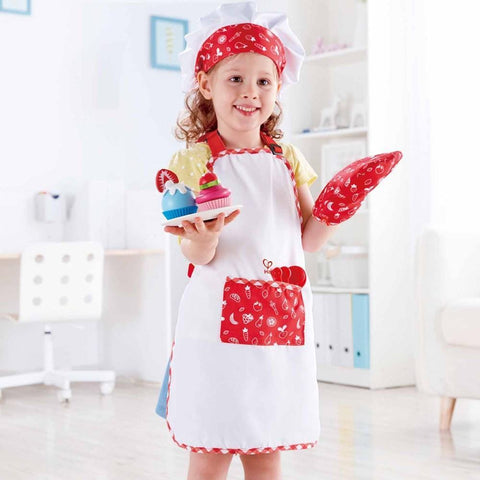 A young girl dressed as a chef, wearing the red and white apron, chef hat, and oven mitt from her Hape Chef Pack by Hape, is smiling and holding two decorated cupcakes. She stands in a bright room with white furniture and decor in the background.