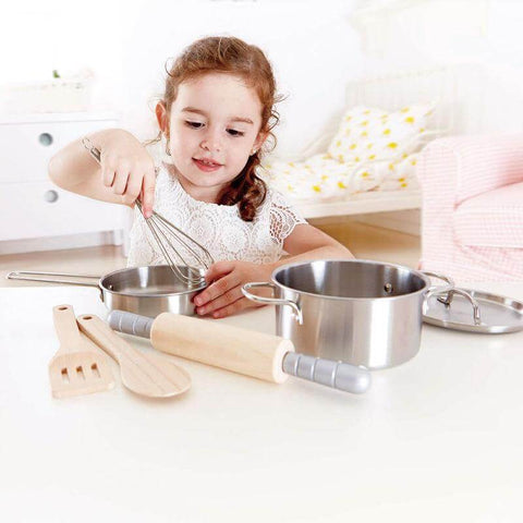 A young child with curly hair, wearing a white shirt, is playing with the Hape Chef's Cooking Set in a bright room. On the table in front of her are a rolling pin, wooden spoons, and another pot with a lid from Hape. Perhaps this future chef is already dreaming of preparing her first healthy meal. In the background, there is a pastel-colored sofa and dresser.