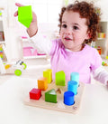 A young child with curly hair is playing with the Hape Color and Shape Sorter. The child is holding a green wooden cube from the Hape set and is about to place it on the wooden board, which features corresponding holes for colorful geometric shapes. The child is smiling and appears engaged in sorting activities, enhancing their color and shape identification skills.