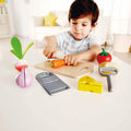 A young child with dark hair, dressed in a light blue striped shirt, sits at a table pretending to slice a toy carrot from the Hape Cooking Essentials set on a wooden cutting board. Scattered across the table are various other toy vegetables and kitchen tools, beautifully depicting the essence of Hape Kitchens.