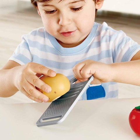 A young child, dressed in a striped shirt, uses a grater on a yellow item while sitting at a table with the Hape Cooking Essentials from Hape. A red toy rests next to the child on what looks like a toy cutting board. The scene is set in a bright room with a light-colored floor.