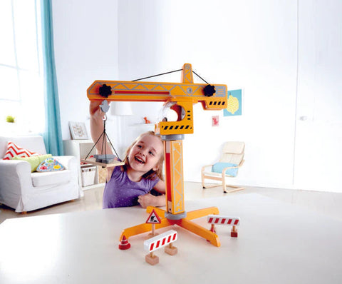 A child with blonde hair wearing a purple shirt is smiling and engaging in pretend play with the Hape Crane Lift on a white table. The room in the background has a white wall, a cushioned chair, a small plant, and colorful pillows on a couch.