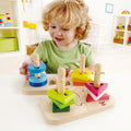 A young child with curly blonde hair and wearing a green t-shirt is playing with the Hape Creative Peg Puzzle, a colorful wooden toy by Hape that enhances their fine motor skills. The puzzle consists of variously shaped blocks on pegs, which the child is happily arranging. The background shows a bright, organized playroom.