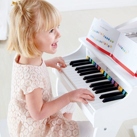 A young girl with blonde hair and a lace dress enthusiastically plays the Hape Deluxe White Grand Piano. The piano, adorned with colorful stickers on the keys, features an open music book resting on the stand above them. She appears excited and engaged, as if composing mini symphonies.