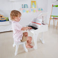 A young child wearing a pink shirt and beige shorts is sitting on a small white chair, playing a miniature Hape Deluxe White Grand Piano in a brightly lit room. In the background, there is a guitar on a white couch, colorful bunting, framed artwork, and an easel.