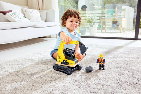 A young child with curly hair sits on a carpeted floor, playing with a Hape Dig 'n Dump Excavator. The child is using the realistic playset to scoop up a small rock-like object. Nearby, there is a small toy construction worker wearing a yellow hard hat and an orange vest. The room is bright with large windows in the background revealing greenery and an outdoor deck.