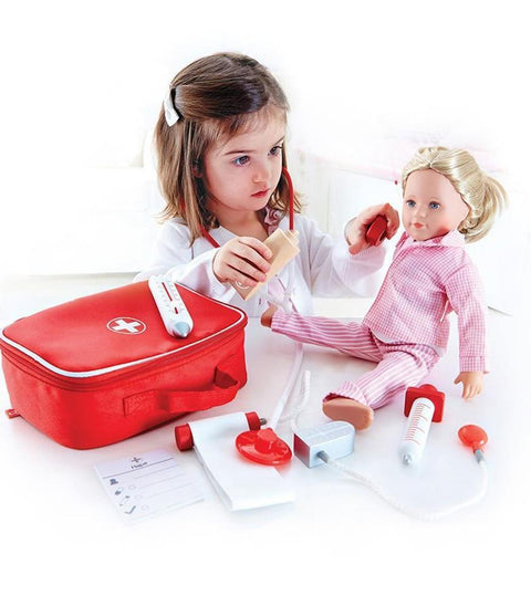 A young girl pretends to be a doctor with Hape's "Doctor On Call" playset, using a toy stethoscope on the blonde-haired, pink-pajama-clad doll. The comprehensive playset includes various medical toy accessories such as a red toy doctor's bag, a notepad, and a thermometer scattered nearby.