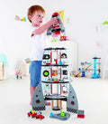 A young child engages with the Hape Four-Stage Rocket Ship toy, ideal for enhancing cognitive skills. The Hape rocket ship features an array of colorful rooms spread across multiple levels, complete with a launch pad at the top. Dressed in a white T-shirt and blue shorts, the child relishes playtime in the brightly lit room.