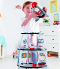 A young boy in a white shirt is joyfully playing with a toy airplane. He stands by the Hape Four-Stage Rocket Ship, a multi-level playset filled with various miniature rooms and space-themed decorations. The room is bright, adorned with children's toys and decor aimed at enhancing cognitive skills.