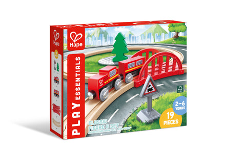 The image shows a box of the Hape Play Essentials Classic Figure 8 Set, a 19-piece wooden train set by Hape designed for children aged 2-6 years. Offering the ultimate railway experience, the box includes a red train, red bridge, track pieces, a tree, and a railroad crossing sign all set against a scenic background.