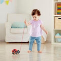 A toddler in a pink top and blue pants is playing indoors, pulling the Hape Pull-along Ladybug with a string. The room is brightly lit, featuring a white armchair, a shelf filled with colorful books, and storage boxes in the background. This delightful children's toy encourages movement and exploration for young ones.