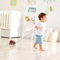 A toddler wearing a white shirt and blue jeans happily pulls a Hape Pull along Ladybug toy across a bright and tidy room with a crib, a white shelf filled with stuffed animals, and various decorations in the background, encouraging movement as they explore their world.