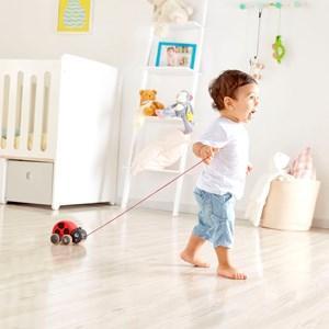 A toddler wearing a white shirt and blue jeans happily pulls a Hape Pull along Ladybug toy across a bright and tidy room with a crib, a white shelf filled with stuffed animals, and various decorations in the background, encouraging movement as they explore their world.