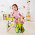 A young girl wearing a pink polka dot tank top and floral pants sits on a green Hape Scoot-Around bike in a brightly lit playroom with white walls. The room is filled with colorful toys, books, and shelves in the background.