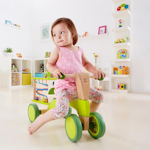 A young girl wearing a pink polka dot tank top and floral pants sits on a green Hape Scoot-Around bike in a brightly lit playroom with white walls. The room is filled with colorful toys, books, and shelves in the background.