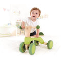 A toddler in a white shirt and blue jeans is riding the Hape Scoot-Around, a green and beige foot-powered bike with four wheels. The child looks happy, building muscle strength indoors in a bright, decorated room with shelves and toys in the background.