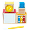 The Hape Stacking Music set by Hape is a wooden children's musical toy featuring colorful percussion instruments. It includes a yellow bell, blue cube, white frame, three small xylophone bars in red, yellow, and blue, and a yellow mallet. All parts are mounted on or stored in wooden blocks for easy stacking.