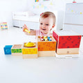 A toddler joyfully plays with the vibrant Hape Stacking Music Set, which includes a variety of musical components such as a xylophone and cymbals. The child is smiling and holding one of the musical pieces, seated at a white table in a bright, modern room.