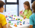 Three children are playing at a table filled with colorful modeling clay figures and molds. A girl in a green shirt is focusing on molding clay, while a boy in yellow and another in blue engage with the Hey Clay 15 pack - Animals and a tablet nearby. The room is well-lit with natural light, making play-based learning enjoyable.