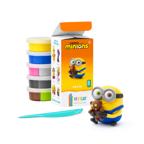 An open box of Hey Clay Single Pack Minions - Bob & Tim from Hey Clay, featuring Bob holding a teddy bear (Tim). The box displays the clay colors and an image of Bob on the front. In front, there are six stacked containers of modeling clay in yellow, white, pink, green, gray, and black—perfect for developing fine motor skills.