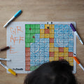 A child is drawing on a Hey Doodle Silicone Activity Mat - 100 Squares (Large) with colored markers. The mat, from Hey Doodle, features color-coded numbered squares and blue spiral patterns, making it perfect as a math teaching aid. The word "GIRAFFE" is written in orange on the mat while various colored markers are placed around it on a wooden surface.