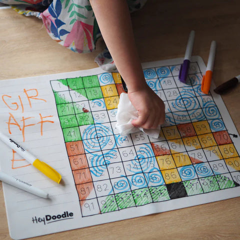 A child uses a cloth to wipe clean a colorful Hey Doodle Silicone Activity Mat - 100 Squares (Large) on the table. The mat, from Hey Doodle, contains various colored and numbered sections, perfect for creative games and drawings made with colored markers. The word "GIRAFFE" is written on the left side of the mat.