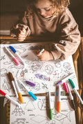 A child with curly hair colors on the Hey Doodle Silicone Playmat - Outer Space (Large) using a marker, surrounded by various colored markers. The mat features spaceship and planet drawings along with geometric shapes waiting to be filled in. The child, dressed in a comfy beige sweater from Hey Doodle, enjoys the open-ended drawing prompts.