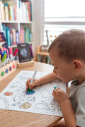 A young child colors a space-themed illustration on the Hey Doodle Silicone Playmat - Outer Space (Large) at a wooden desk. The image includes planets, astronauts, and spaceships. A bookshelf filled with colorful books and a window with blinds are in the background, making it the perfect setting for Hey Doodle mats and open-ended drawing prompts.