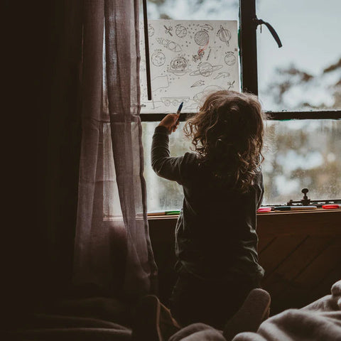 A small child with curly hair stands by a window, eagerly drawing on a Hey Doodle Silicone Playmat - Outer Space (Large) taped to the glass. The window is partially covered by a translucent curtain, allowing some natural light to filter through. The faint outline of outdoor greenery can be seen in the background.