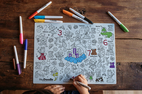 A child colors on a Hey Doodle Silicone Playmat - Sugar & Spice (Large), featuring various whimsical characters, including princesses, dragons, and unicorns. Colored markers are scattered around the reusable silicone mat on a wooden table. The placemat includes numbers and a colorful central scene with a castle on a cloud.