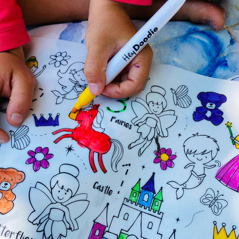 A child's hands are shown using coloring markers on a Hey Doodle Silicone Playmat - Sugar & Spice (Large) featuring fairies, a castle, flowers, teddy bears, and other whimsical designs. The child is using a yellow marker to color a portion of the picture. The image captures the creative process of designing on this reusable silicone mat by Hey Doodle.