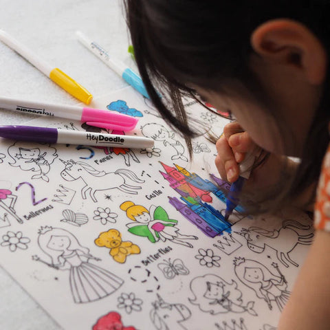 A child joyfully colors a Hey Doodle Silicone Playmat - Sugar & Spice (Large) with printed images of castles, fairies, and unicorns. Using a blue marker to fill in the details, they have other colored markers in yellow, pink, purple, and green scattered on the table. This reusable silicone playmat from Hey Doodle makes creativity endless.