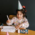 A young child wearing a party hat and a floral-patterned sweater is seated at a table, coloring in a sketchbook with markers. Colorful paper cups are scattered around her on the Hey Doodle Silicone Playmat - Sugar & Spice (Mini). The background is dark, making the child the focal point of the image.