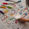 A child erases the Hey Doodle Silicone Playmat - Toot Toot Honk (Large), which showcases colorful illustrations of vehicles like a school bus, helicopter, truck, and tractor, using a cloth. Several colored markers are scattered around the reusable silicone mat from Hey Doodle.