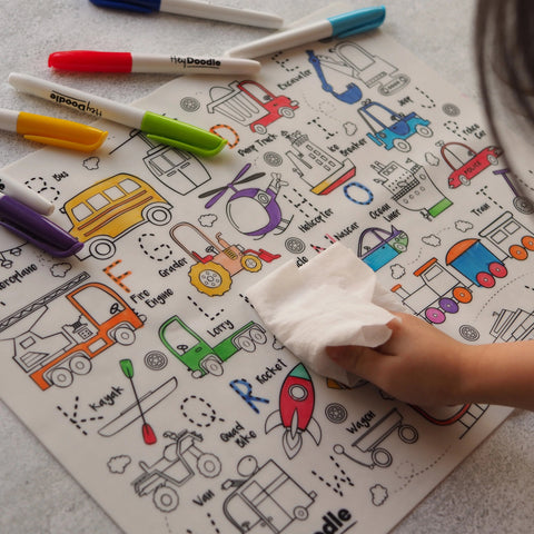 A child erases the Hey Doodle Silicone Playmat - Toot Toot Honk (Large), which showcases colorful illustrations of vehicles like a school bus, helicopter, truck, and tractor, using a cloth. Several colored markers are scattered around the reusable silicone mat from Hey Doodle.