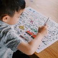 A young child is seated at a wooden table, drawing on the Hey Doodle Silicone Playmat - Toot Toot Honk (Large) filled with vehicle illustrations and labels. The child holds a white marker, focused on coloring in a red train. The background is out of focus, showcasing the engaging Hey Doodle mats designed for learning alphabets through fun activities.