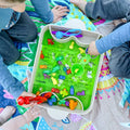 Two children sit on a Hey Doodle Silicone Playmat - Toot Toot Honk (Mini) by Hey Doodle, playing with a sensory bin filled with green slime and various plastic toys, including animals and tools. The bin, adorned with a white border and some doodles, has one child holding red tongs while the other uses a green scoop to discover hidden treasures like washable markers.