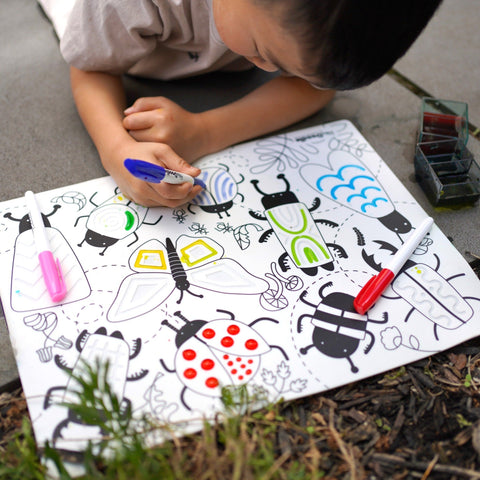 A child is lying on a Hey Doodle Silicone Sensory Mat - Busy Bugs (Large), coloring a large sheet filled with black-and-white illustrations using colored markers. The image shows partially colored butterflies, ladybugs, and other insects from the Hey Doodle brand, enhancing the child's fine motor skills. A set of colored markers is nearby.