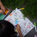 Children coloring on a Hey Doodle Silicone Sensory Mat - Wild Digits (Large) outdoors, surrounded by greenery. The mat features various animals and numbers. Colored markers and an open paint set are visible, enhancing fine motor skills. A child’s hand is seen coloring with a blue marker.