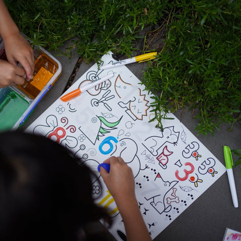 Children coloring on a Hey Doodle Silicone Sensory Mat - Wild Digits (Large) outdoors, surrounded by greenery. The mat features various animals and numbers. Colored markers and an open paint set are visible, enhancing fine motor skills. A child’s hand is seen coloring with a blue marker.