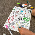 A child's hand colors on a Hey Doodle Silicone Sensory Mat - Wild Digits (Large) featuring animals and numbers using various colored markers, honing their fine motor skills. The mat lies on a textured surface, surrounded by markers with caps off—green, yellow, red, and blue. The child's arm is partially visible.