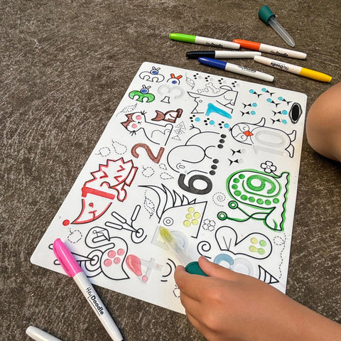 A child's hand colors on a Hey Doodle Silicone Sensory Mat - Wild Digits (Large) featuring animals and numbers using various colored markers, honing their fine motor skills. The mat lies on a textured surface, surrounded by markers with caps off—green, yellow, red, and blue. The child's arm is partially visible.