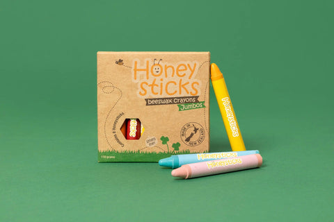 A box of Honeysticks Beeswax Crayons Jumbo (8pk) by Honeysticks is positioned against a green background, alongside three crayons: yellow, blue, and pink. The box showcases eco-friendly design features with a small window displaying one non-toxic crayon inside.